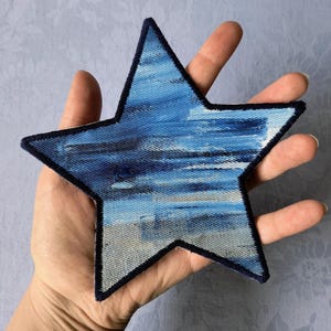 Puede incluir: Un parche azul en forma de estrella con un borde azul oscuro. El parche presenta un diseño texturizado con tonos de azul y gris, que se asemeja a una pintura de acuarela. El parche se sostiene en una mano.