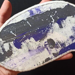 Może przedstawiać: Abstrakcyjna naszywka z białym, szytym obramowaniem. Naszywka ma wzór z czarnymi, fioletowymi i białymi plamami farby. Naszywka jest trzymana w dłoni.