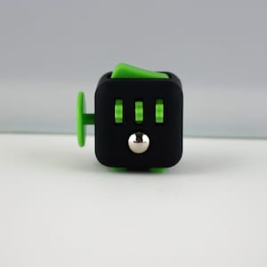 Pode incluir: Um cubo fidget preto com detalhes verdes. O cubo apresenta um interruptor basculante verde, três botões de clique verdes, uma bola de metal prateado e um joystick verde. Projetado para alívio do estresse e jogo sensorial.