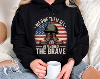 May include: Black hoodie with a graphic design featuring a military helmet, boots, and dog tags in front of an American flag. The text reads "WE OWE THEM ALL" and "WE REMEMBER THE BRAVE". The hoodie is worn by a person holding a white mug.