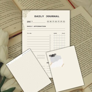 May include: A daily journal page with a header, date line, days of the week, a daily affirmation section, and a to-do list with work and play columns. Two smaller note cards are layered on top, one with a grid pattern and one with a moth graphic.
