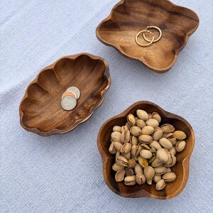 May include: Three wooden, flower-shaped bowls on a light blue surface. One bowl holds pistachios, another has coins, and the third contains gold hoop earrings. The bowls are made of a dark wood with a natural grain.