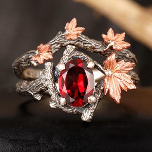 Könnte beinhalten: Silberner Ring mit einem großen, ovalen, tiefroten Edelstein. Der Ring hat ein von der Natur inspiriertes Design mit Ästen und kupferfarbenen Ahornblättern. Die Blätter und Äste umschließen den Edelstein.