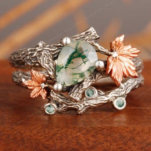 Könnte beinhalten: Ein silberner Ring mit einem birnenförmigen grünen Edelstein, akzentuiert durch kupferfarbene Ahornblätter und kleine grüne Steine. Das Ringband ist als Baumzweige gestaltet und schafft eine von der Natur inspirierte Ästhetik. Der Ring ist ein einzigartiges Schmuckstück.