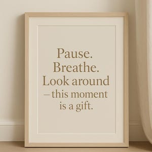 May include: Framed print featuring the text "Pause. Breathe. Look around - this moment is a gift." in a brown font. The print is in a white mount and a light wood frame. The background is a neutral colour.