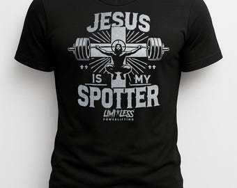 Puede incluir: Camiseta negra con un diseño gráfico plateado. El diseño presenta el texto "JESUS IS MY SPOTTER" y una imagen de Jesús levantando pesas en una cruz. La camiseta también tiene el texto "LIMITLESS POWERLIFTING".
