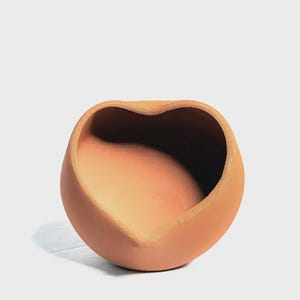 Puede incluir: Un cuenco de terracota en forma de corazón con base redondeada. El cuenco tiene una superficie lisa y sin esmaltar y un tono cálido y terroso. El interior es un tono más oscuro del mismo color. El cuenco probablemente se utiliza con fines decorativos.