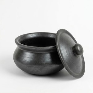 以下が含まれることがあります： 黒色の手作りの土鍋と蓋。 蓋はわずかに開いており、鍋の暗い内部が見えます。 鍋は丸みを帯びた本体と広い縁があり、スープやシチューの提供に最適です。