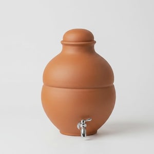 May include: A terracotta water dispenser with a rounded body and a matching lid. The vessel features a smooth, matte finish in a warm reddish-brown clay tone. A polished silver-colored metal spigot is attached to the lower front section.