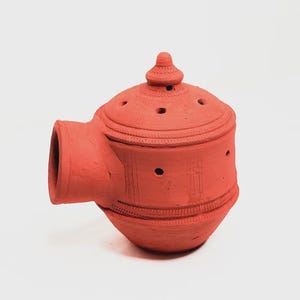 May include: A terracotta-colored incense burner with a rounded base, a lid with small holes, and a side spout. The burner has decorative etched lines and a small, conical finial on top. The overall shape is reminiscent of a teapot.