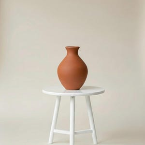 Könnte beinhalten: Eine terrakottafarbene Vase mit rundem Körper und schmalem Hals steht auf einem kleinen, weißen Hocker mit runder Oberseite. Die Vase hat eine glatte Oberfläche und ein schlichtes Design.