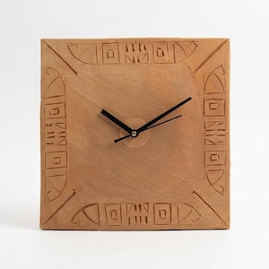 Op de afbeelding: Vierkante, terracotta-kleurige klok met zwarte wijzers. De wijzerplaat heeft gesneden geometrische patronen langs de randen. De wijzers zijn zwart en geven de tijd aan. De klok is een decoratief item.