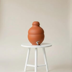 May include: A terracotta water dispenser with a silver spigot and a matching lid. The dispenser is placed on a small, round, white wooden stool. The background is a solid, neutral color.