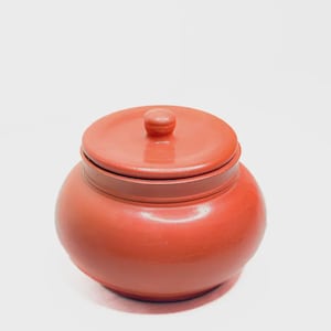 May include: A small, round, terracotta-colored ceramic jar with a matching lid. The lid has a small knob on top for easy removal. The jar has a smooth, glossy finish and sits on a white surface.