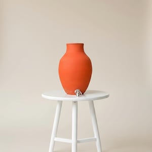 Puede incluir: Un dispensador de agua de terracota naranja con un grifo plateado se encuentra sobre una pequeña mesa redonda blanca con tres patas. El dispensador tiene un cuerpo redondeado y un cuello estrecho. El fondo es de color neutro.