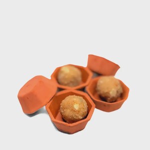 Puede incluir: Vista aérea de varios cuencos naranjas de forma geométrica, cada uno con un alimento redondo de color marrón dorado. La comida parece ser una pequeña bola texturizada, posiblemente un postre o un aperitivo. Los cuencos están colocados sobre un fondo claro.