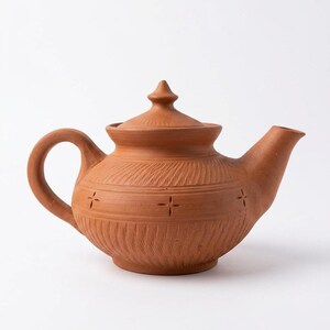 Puede incluir: Una tetera de terracota con tapa y asa. La tetera tiene un cuerpo redondeado con líneas grabadas y cortes en forma de cruz. El pico está en ángulo y el asa es un bucle curvado. La tetera es de color marrón cálido y terroso.