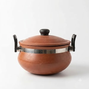 May include: A round terracotta cooking pot with a matching lid, featuring a smooth, reddish-brown clay finish. The pot is encircled by a wide, polished silver-tone metal band that connects two black plastic side handles and a black lid knob.