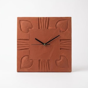 May include: A square terracotta wall clock with a matte finish. The clock face features a central square panel surrounded by four leaf-shaped indentations at the corners and linear carved patterns on each side. It has two thin black hands.
