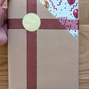 K&ouml;nnte beinhalten: Ein rechteckiger, in braunes Papier eingewickelter Gegenstand mit einem vertikalen dunkelbraunen Band und einem goldenen runden Siegel. Eine Ecke der Verpackung ist offen und gibt den Blick auf eine wei&szlig;e Karte mit roten und goldenen Luftballons, Konfetti und dem Text Happy Birthday frei.