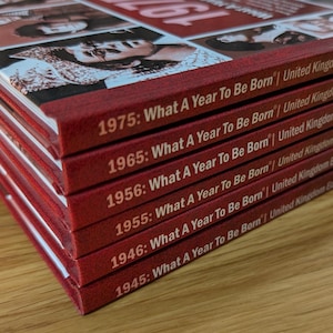K&ouml;nnte beinhalten: Ein Stapel aus sechs roten Hardcover-B&uuml;chern mit wei&szlig;em Text auf den Buchr&uuml;cken. Jeder Buchr&uuml;cken zeigt das Jahr gefolgt vom Titel What A Year To Be Born | United Kingdom. Die aufgef&uuml;hrten Jahre sind 1975, 1965, 1956, 1955, 1946 und 1945.