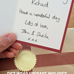 K&ouml;nnte beinhalten: Eine wei&szlig;e Gru&szlig;karte mit dunkelrotem Rand und handgeschriebenem Text in schwarzer Tinte: Happy 70th Birthday Richard, Have a wonderful day, Lots of love, John & Sheila, xxx. Die Karte liegt auf einem braun verpackten Geschenk.