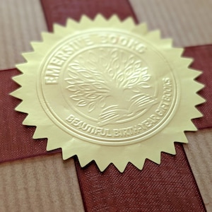 K&ouml;nnte beinhalten: Ein braunes Geschenkpapierpaket mit einem dunkelroten Band in Kreuzform, verziert mit einem runden goldenen Foliensiegel. Das Siegel zeigt ein gepr&auml;gtes Baummotiv und den Text EMERSIVE BOOKS BEAUTIFUL BIRTH-YEAR GIFT BOOKS.