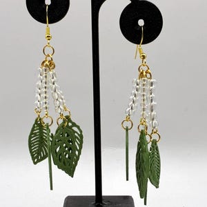 May include: Dangle earrings with gold-coloured hooks, clear beads, and green leaf charms. The earrings are displayed on a black stand. Each earring has three green leaf-shaped charms and gold-coloured accents. The background is white.