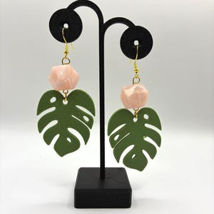 May include: A pair of green Monstera leaf earrings with a pink geometric bead and gold-coloured hooks. The earrings are displayed on a black jewellery stand against a white background. The earrings are a tropical theme.