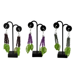 May include: Three pairs of dangle earrings on black display stands. Each earring has a beaded strand in brown, purple, or grey, with green leaf charms. The earrings are displayed against a white background.