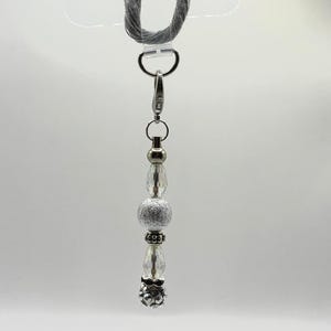 May include: A silver beaded keychain with a lobster clasp and a gray cord loop. The keychain features clear, faceted beads, a silver glitter bead, and decorative silver accents. The bottom bead has a floral design.