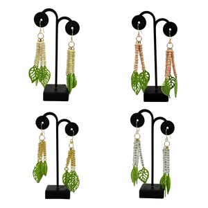 May include: Four pairs of dangle earrings on black display stands. Each pair features a unique combination of beads and green leaf-shaped charms. The earrings vary in colour, including cream, pink, gold, and clear beads.