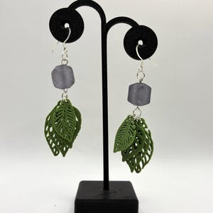 May include: Dangle earrings with a grey, faceted bead and green leaf charms. The earrings hang from silver-coloured hooks. The leaves have a detailed, openwork design. The earrings are displayed on a black stand.