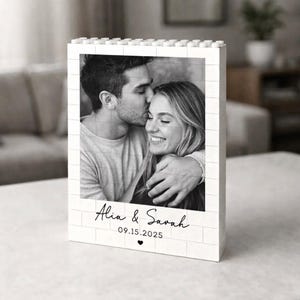 Puede incluir: Fotografía en blanco y negro de una pareja en un marco blanco con diseño de ladrillos. El marco muestra los nombres "Alia & Sarah", la fecha "09.15.2025" y un corazón. La imagen muestra a un hombre besando a una mujer en la frente.