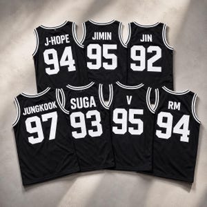 Peut inclure: Sept maillots de basket-ball noirs avec des bordures blanches autour du col et des emmanchures. Chaque maillot affiche un nom et un numéro blancs au dos : J-HOPE 94, JIMIN 95, JIN 92, JUNGKOOK 97, SUGA 93, V 95 et RM 94, en police blanche.