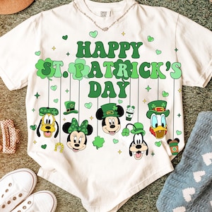Puede incluir: Camiseta blanca con un diseño del Día de San Patricio. La camiseta presenta el texto "Happy St. Patrick's Day" en verde, junto con caras de personajes de dibujos animados con sombreros verdes y tréboles. Se ve un collar.