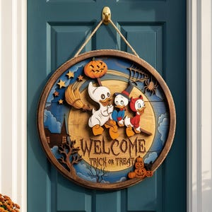 May include: A round wooden door sign featuring three cartoon ducks in Halloween costumes, a broom, a jack-o'-lantern, and a spider web. The sign displays the text "WELCOME" and "TRICK OR TREAT" in a dark font against a yellow moon background.