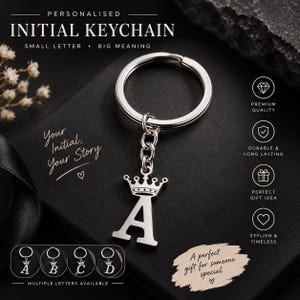 Pode incluir: Um porta-chaves de metal prateado com um pendente em forma de letra maiúscula A encimado por uma pequena coroa. O pendente está preso a uma argola circular por uma corrente curta. O texto na imagem inclui Personalised Initial Keychain e Your Initial.