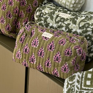 Puede incluir: Un surtido de bolsas de viaje y neceseres con estampado de cachemira en verde oliva y rosa. También hay una bolsa cilíndrica a cuadros verdes y blancos y una bolsa verde con estampado floral. Las bolsas están apiladas sobre una superficie verde oscuro.