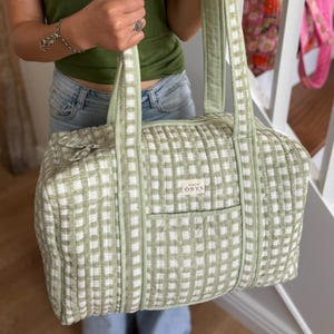 May include: A light green and white checked quilted duffel bag with matching handles. The bag has a rectangular shape with a small front pocket and the brand name "ORYS" on a tag. The bag is held by a person.