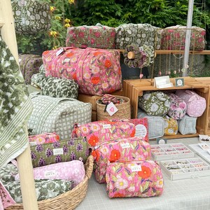 May include: A market stall showcasing a variety of floral-patterned bags and accessories in pink, green, and white. The bags include duffel bags and cosmetic pouches. Some items are displayed in a woven basket and on wooden shelves.
