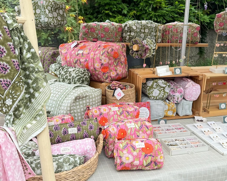 May include: A market stall showcasing a variety of floral-patterned bags and accessories in pink, green, and white. The bags include duffel bags and cosmetic pouches. Some items are displayed in a woven basket and on wooden shelves.