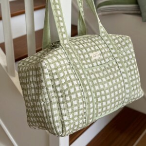 May include: A green and white checked duffel bag with matching handles. The bag is rectangular and has a small front pocket. The bag is suspended from a white banister. The bag has the word "ORYS" on the front.