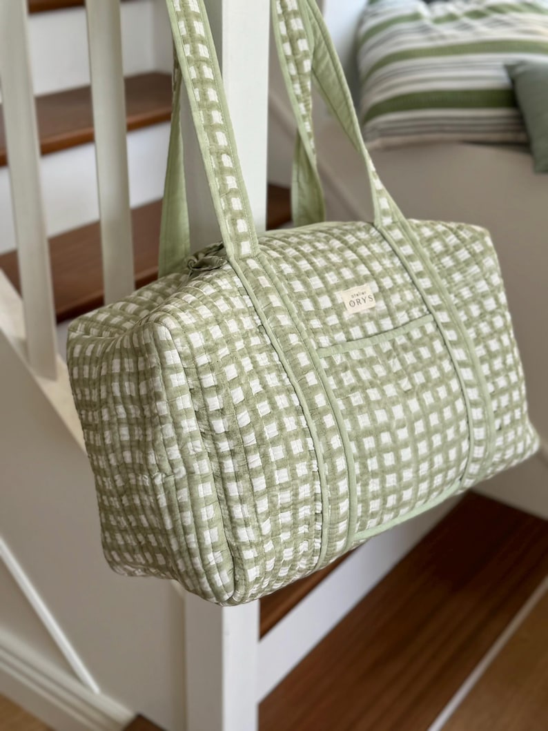 May include: A green and white checked duffel bag with matching handles. The bag is rectangular and has a small front pocket. The bag is suspended from a white banister. The bag has the word "ORYS" on the front.