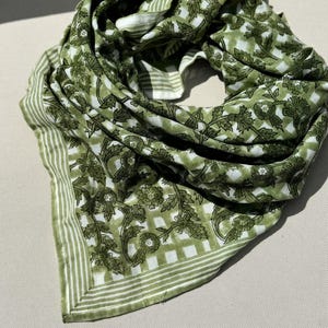 Peut inclure: Écharpe à motifs verts et blancs. L'écharpe présente un motif floral et à carreaux avec une bordure rayée. L'écharpe est faite d'un tissu léger et est drapée en forme circulaire.
