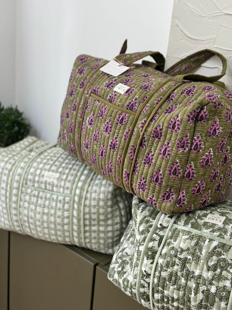 May include: Three quilted travel bags in shades of green. The top bag is olive green with a pink paisley pattern. The middle bag is light green with a grid pattern. The bottom bag is light green with a floral pattern. Each bag has a tag that says "ORYS".