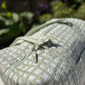 May include: A light green and white checked duffel bag with a zip closure. The bag has a quilted texture and a matching handle. The bag is made of a soft, lightweight material.