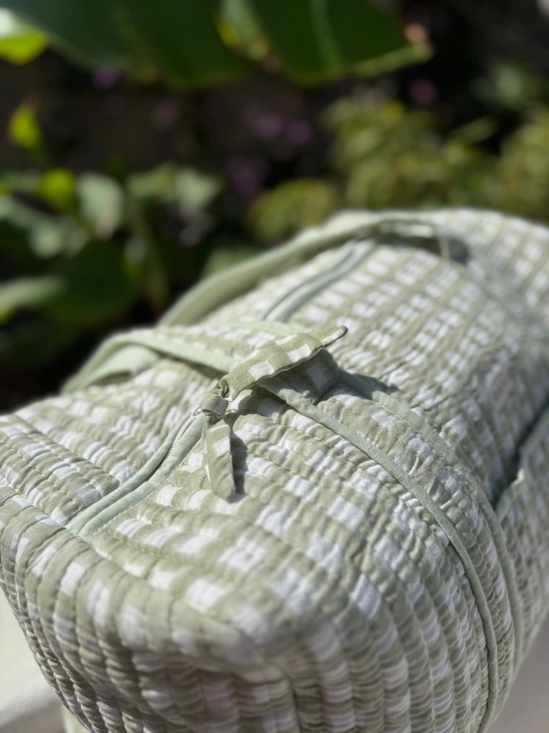 May include: A light green and white checked duffel bag with a zip closure. The bag has a quilted texture and a matching handle. The bag is made of a soft, lightweight material.