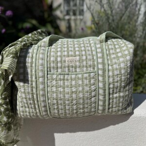 May include: A light green and white checkered duffel bag with a matching strap. The bag has a rectangular shape with a small pocket on the front and the text "atelier ORYS" on a small tag. The bag is made of a quilted fabric.