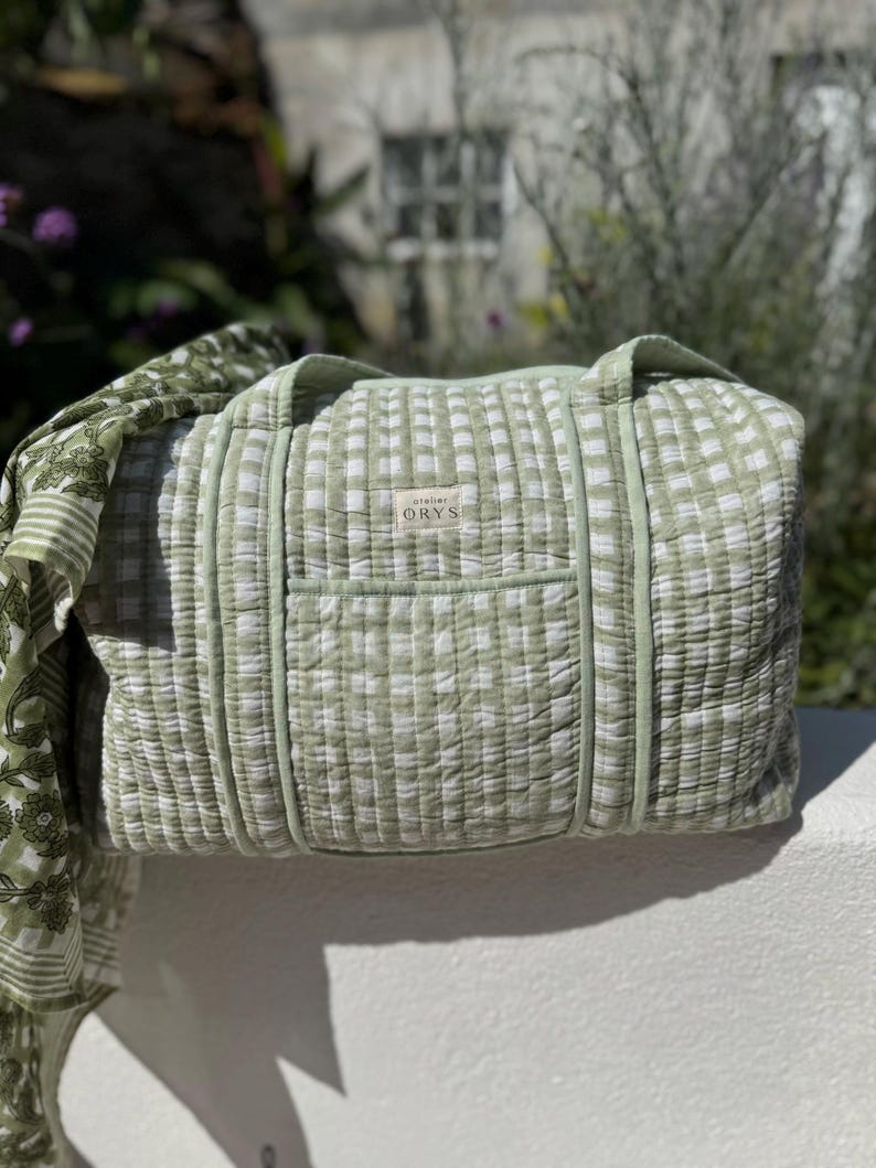 May include: A light green and white checkered duffel bag with a matching strap. The bag has a rectangular shape with a small pocket on the front and the text "atelier ORYS" on a small tag. The bag is made of a quilted fabric.